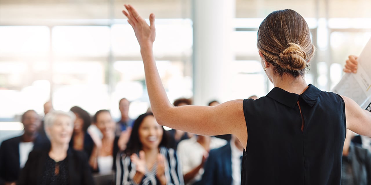Photo of a woman with her arms raised in the air standing in front of a room of people that are clapping.